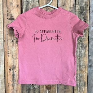 Könnte beinhalten: Ein pinkfarbenes T-Shirt mit dem Text "So apparently, I'm Dramatic" in Schwarz.