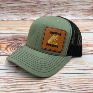 May include: A green and black trucker hat with a brown leather patch. The patch features a corn graphic and the text "AMERICA NEEDS FARMERS". The hat has a curved brim and a mesh back.