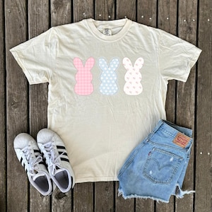 May include: A cream colored t-shirt with three pastel bunny silhouettes. The bunnies are patterned with gingham, floral, and polka dots. The shirt is laying on a wooden surface with a pair of white sneakers and denim shorts.