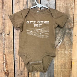 Op de afbeelding: Een bruine baby romper met korte mouwen met een witte afbeelding van een boerderijsc&egrave;ne en de tekst "Cattle Checking Buddy".