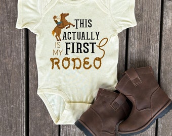 May include: A cream-colored baby onesie with a brown cowboy riding a brown horse. The onesie has black text that reads "This is my first rodeo" with a brown rope design around the words.