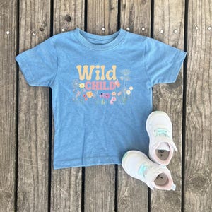Peut inclure: Un t-shirt bleu clair avec le texte "Wild Child" en lettres jaunes et roses, entouré de fleurs colorées. Le t-shirt est sur une surface en bois avec deux baskets blanches avec des accents bleu clair.