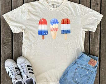 May include: Cream-colored t-shirt featuring a watercolor design of three patriotic ice popsicles. The shirt is paired with light-wash denim shorts and white sneakers with black stripes. The image is set on a wooden surface.