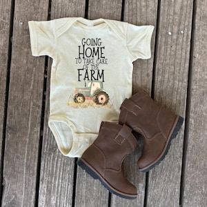 May include: A light beige baby onesie with the text "Going Home to Take Care of the Farm" and a tractor graphic. Paired with brown leather baby boots.
