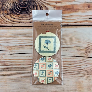 May include: Two car coasters with floral designs are packaged in a clear plastic sleeve. One coaster features a blue flower in a decorative frame, while the other has a repeating pattern of flowers in squares. The text "Car Coaster" is visible.