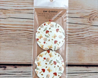 May include: Two car coasters with a white background and a pattern of red mushrooms, butterflies, and leaves. The coasters are packaged in a clear plastic bag with a cardboard backing. Text on the backing reads "Car Coaster".