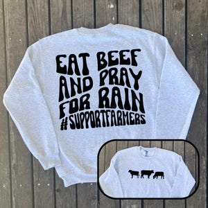 May include: A light grey sweatshirt with the slogan "EAT BEEF AND PRAY FOR RAIN #SUPPORTFARMERS" in black. A small image of three black cow silhouettes is in the lower right corner. The sweatshirt is displayed on a wooden surface.