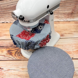 May include: A white KitchenAid Classic stand mixer with a patchwork fabric cover in red, white, and blue patterns. A matching gray cover sits below. The mixer is on a wooden surface.