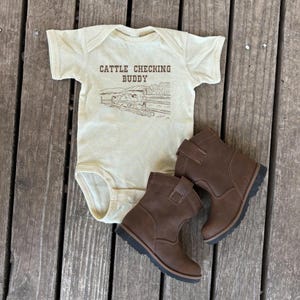 Op de afbeelding: Een cr&egrave;mekleurige baby romper met de tekst "Cattle Checking Buddy" en een tekening van een boerensc&egrave;ne. Naast de romper ligt een paar bruine leren laarzen.