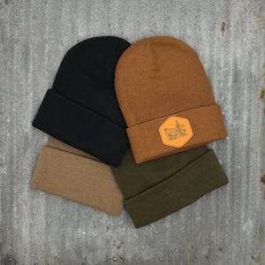 May include: Three knit beanies in black, brown, and green. The brown beanie has a leather patch with a desert scene.