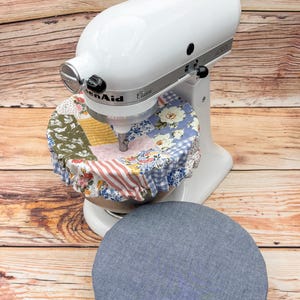 May include: A white KitchenAid Classic stand mixer with a colorful patchwork bowl cover and a separate blue cover. The bowl cover features floral and geometric patterns in various colors. The mixer is on a wooden surface.