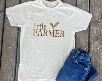 Puede incluir: Una camiseta de color crema con texto marrón que dice "little FARMER" y un pequeño gráfico de trigo marrón.