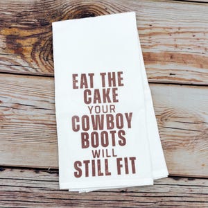 May include: White kitchen towel with the text "EAT THE CAKE YOUR COWBOY BOOTS WILL STILL FIT" in brown lettering. The towel is folded and resting on a wooden surface.