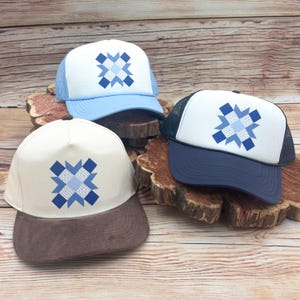 May include: Three trucker hats with a geometric design. The hats have a white front panel with a blue and white starburst pattern. One hat has a light blue mesh back and brim, another has a navy blue mesh back and brim, and the third has a tan front panel and a brown brim.