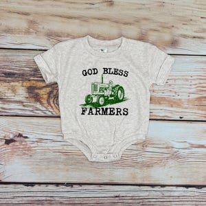 May include: A light grey baby bodysuit with short sleeves. It has a green tractor graphic and the text "GOD BLESS FARMERS" in black. The bodysuit is made of soft material.