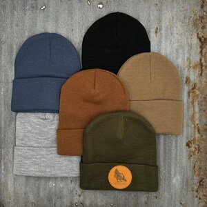 May include: Six different colored knit beanies. The beanies are all folded in half and stacked on top of each other. The beanie on top is olive green with a brown patch that says "Wildcraft".
