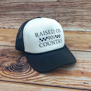 May include: A black and white trucker hat with the text "RAISED ON 90S COUNTRY". The hat has a white front panel, a black brim, and a black mesh back. The text is in a bold, sans-serif font.