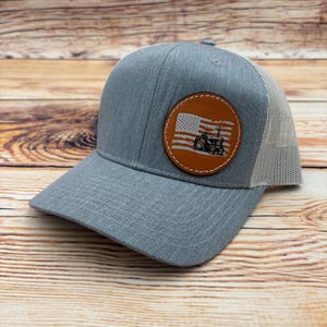 May include: A gray trucker hat with a mesh back and a curved brim. The front features a round, brown leather patch with an American flag design and a tractor silhouette. The hat is made of a durable fabric and has a snapback closure.