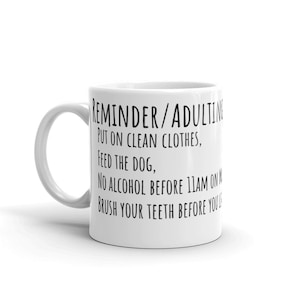 May include: White ceramic mug with black text that reads "REMINDER/ADULTING PUT ON CLEAN CLOTHES, FEED THE DOG, NO ALCOHOL BEFORE 11AM ON, BRUSH YOUR TEETH BEFORE YOU"