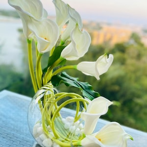 May include: A white calla lily flower arrangement in a clear glass vase with white stones. The vase has a unique design with green stems wrapped around it.