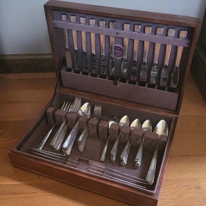 Puede incluir: Un cofre de madera para cubiertos con tapa abisagrada, abierto para revelar un juego completo de utensilios de plata. El juego incluye tenedores, cuchillos y cucharas, cuidadosamente dispuestos en un interior forrado de terciopelo.
