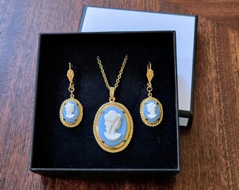 May include: A gold necklace and earrings set with blue cameo pendants featuring a woman's profile. The jewelry is displayed in a black velvet box.