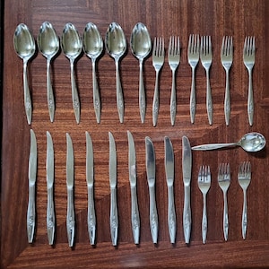 May include: A collection of stainless steel silverware, including spoons, forks, and knives, arranged on a dark wooden surface. The silverware has a floral design on the handles. The set includes various sizes of spoons, forks, and knives.