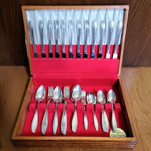 Peut inclure: Un ensemble de couverts anciens présenté dans un coffret en bois avec un intérieur en velours rouge. L'ensemble comprend des couteaux, des fourchettes, des cuillères et des ustensiles de service. Les couverts ont une finition argentée brillante. La boîte est ouverte.
