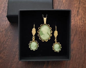 May include: A gold necklace and earrings set with green cameo pendants featuring a woman's profile. The jewelry is displayed in a black velvet box.