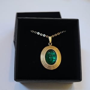 May include: A gold chain necklace with a green glass scarab beetle pendant. The pendant is set in a gold oval frame with concentric circles.