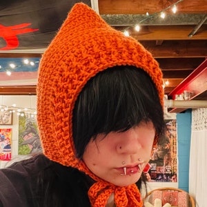 May include: A person wearing a bright orange crocheted hat with a pointed top and a long tie. The hat is pulled down over their forehead, and they have black hair. The person is wearing a black shirt and has multiple facial piercings.