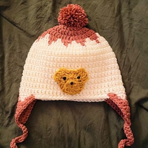Könnte beinhalten: Weißer und rosa gestrickter Hut mit einem braunen Bärenkopf und einem rosa Bommel oben.