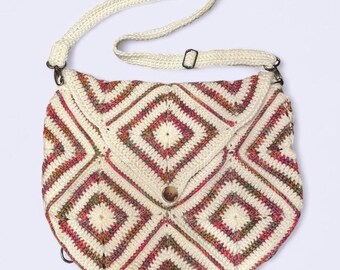 Könnte beinhalten: Cremefarbene Häkeltasche mit geometrischem Rautenmuster in Rot-, Rosa- und Brauntönen. Die Tasche hat einen Klappenverschluss mit einem Holzknopf und einen cremefarbenen Riemen. Die Tasche ist etwa 25 cm breit.