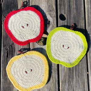 Puede incluir: Tres salvamanteles hechos a mano en ganchillo con forma de manzana. Cada salvamanteles presenta un centro blanco con detalles de semillas marrones y un borde de color: rojo, verde y amarillo. Los salvamanteles están colocados sobre una superficie de madera envejecida.