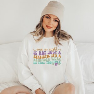 Pode incluir: Camisola branca com o texto "one in eight is not just a statistic it's a reminder we're in this together" em letras coloridas de estilo retro.