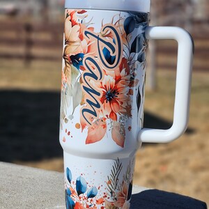 Peut inclure: Un gobelet isotherme blanc avec une poign&eacute;e, orn&eacute; d'un motif floral aux tons orange, bleu et vert. Le nom "Pam" est &eacute;crit en cursive sur le c&ocirc;t&eacute;. Le gobelet a un couvercle et une poign&eacute;e blancs.
