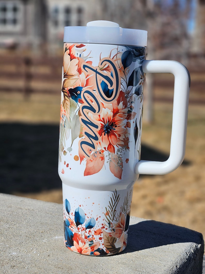 Peut inclure: Un gobelet isotherme blanc avec une poign&eacute;e, orn&eacute; d'un motif floral aux tons orange, bleu et vert. Le nom "Pam" est &eacute;crit en cursive sur le c&ocirc;t&eacute;. Le gobelet a un couvercle et une poign&eacute;e blancs.