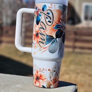 Peut inclure: Un gobelet blanc avec une grande poign&eacute;e, orn&eacute; d'un motif floral aux tons orange, bleu et gris. Le nom "Pam" est &eacute;crit en cursive. Le gobelet est surmont&eacute; d'un couvercle blanc.