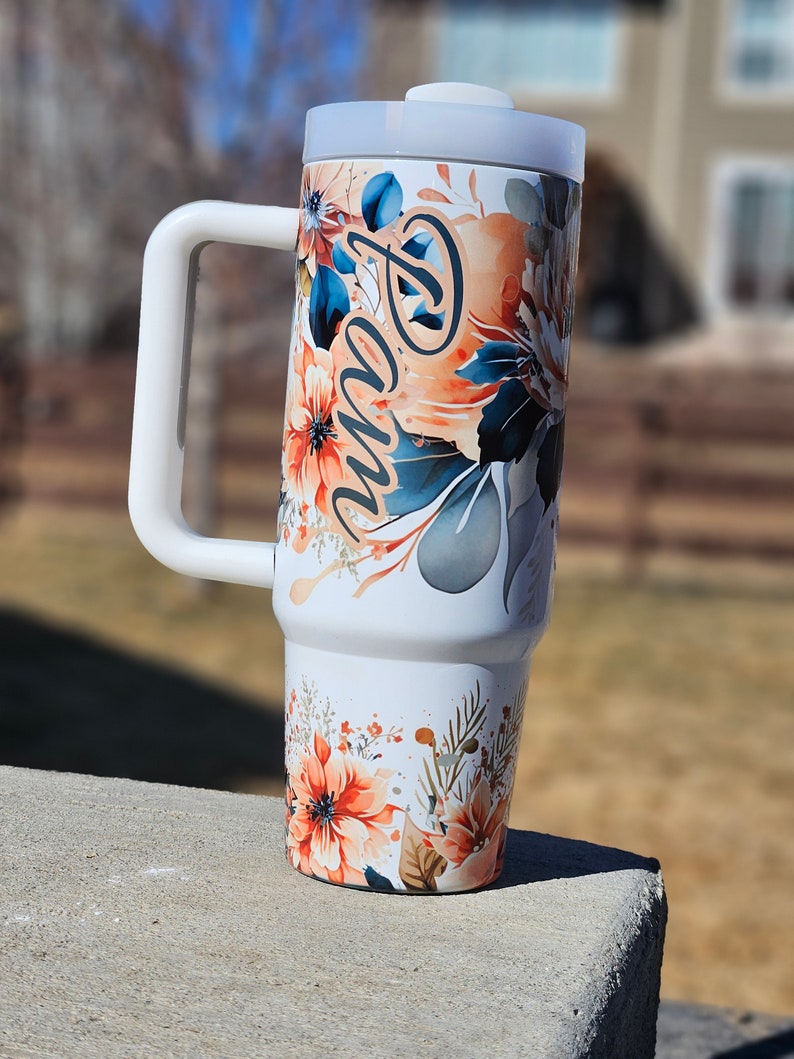 Peut inclure: Un gobelet blanc avec une grande poign&eacute;e, orn&eacute; d'un motif floral aux tons orange, bleu et gris. Le nom "Pam" est &eacute;crit en cursive. Le gobelet est surmont&eacute; d'un couvercle blanc.