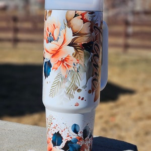 Peut inclure: Gobelet de voyage blanc avec un motif floral dans les tons orange, marron et bleu. Le gobelet a une poign&eacute;e et un couvercle gris clair. Le motif floral comprend de grandes fleurs et des feuilles. Le mot "Grace" est &eacute;crit sur le c&ocirc;t&eacute;.