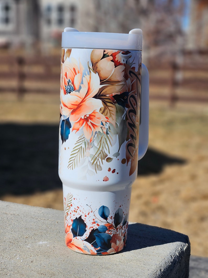 Peut inclure: Gobelet de voyage blanc avec un motif floral dans les tons orange, marron et bleu. Le gobelet a une poign&eacute;e et un couvercle gris clair. Le motif floral comprend de grandes fleurs et des feuilles. Le mot "Grace" est &eacute;crit sur le c&ocirc;t&eacute;.