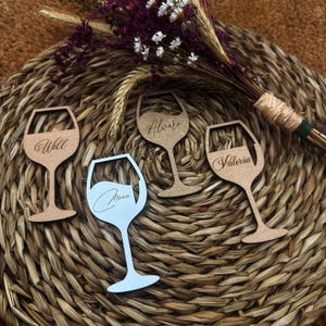 Puede incluir: Cuatro tarjetas de lugar de madera con forma de copa de vino con nombres grabados: Will, Alvaro, Valeria y Maria. Las tarjetas de lugar son en tonos marrón y plateado. Las tarjetas de lugar están dispuestas sobre un mantel individual redondo tejido con flores secas.