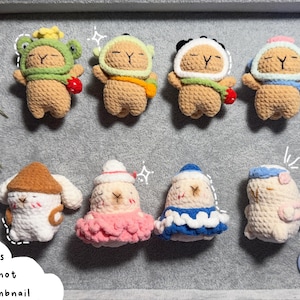 May include: A collection of handmade crochet capybaras in various outfits, including a frog hat, a panda hat, and a mushroom hat. The capybaras are tan with white accents and are displayed on a gray background. The text "NEW! More capybaras in variations not shown in thumbnail" is also visible.