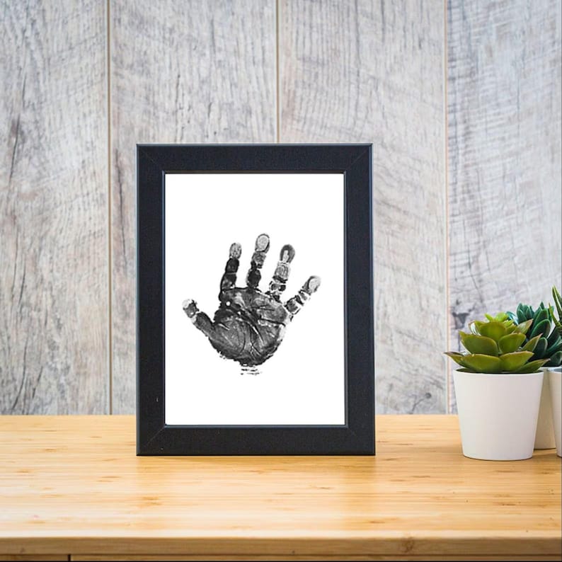 May include: A black framed print of a baby's handprint in black ink on a white background.