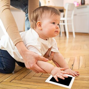 May include: A baby in a white shirt reaches for a black ink pad on a white frame, while being held by an adult.
