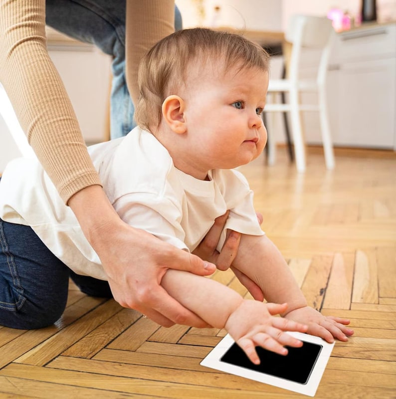 May include: A baby in a white shirt reaches for a black ink pad on a white frame, while being held by an adult.