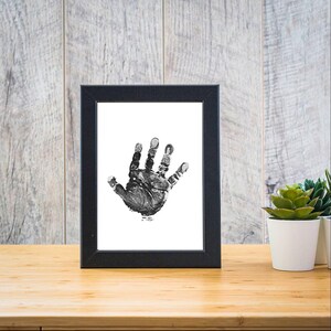 May include: A black framed print of a baby's handprint in black ink on a white background.