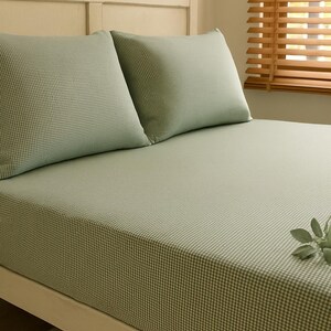 Peut inclure: Un lit avec un drap-housse à carreaux vichy vert clair et deux taies d'oreiller assorties. Le cadre de lit est blanc et la tête de lit est beige clair. Une brindille de verdure repose sur le lit. La pièce a un store en bois.