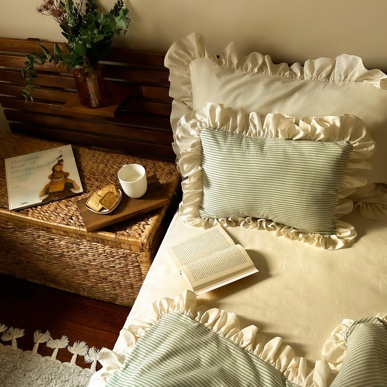 May include: A white bed with a green and white striped pillow and a white pillow with ruffles. A book is on the bed and a small wooden tray with cookies and a cup is on a wicker table next to the bed.