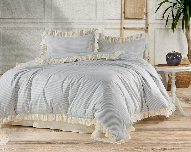 May include: A white and gray striped duvet cover with a ruffled edge. The duvet cover is on a bed with two white and gray striped pillowcases with ruffled edges.
